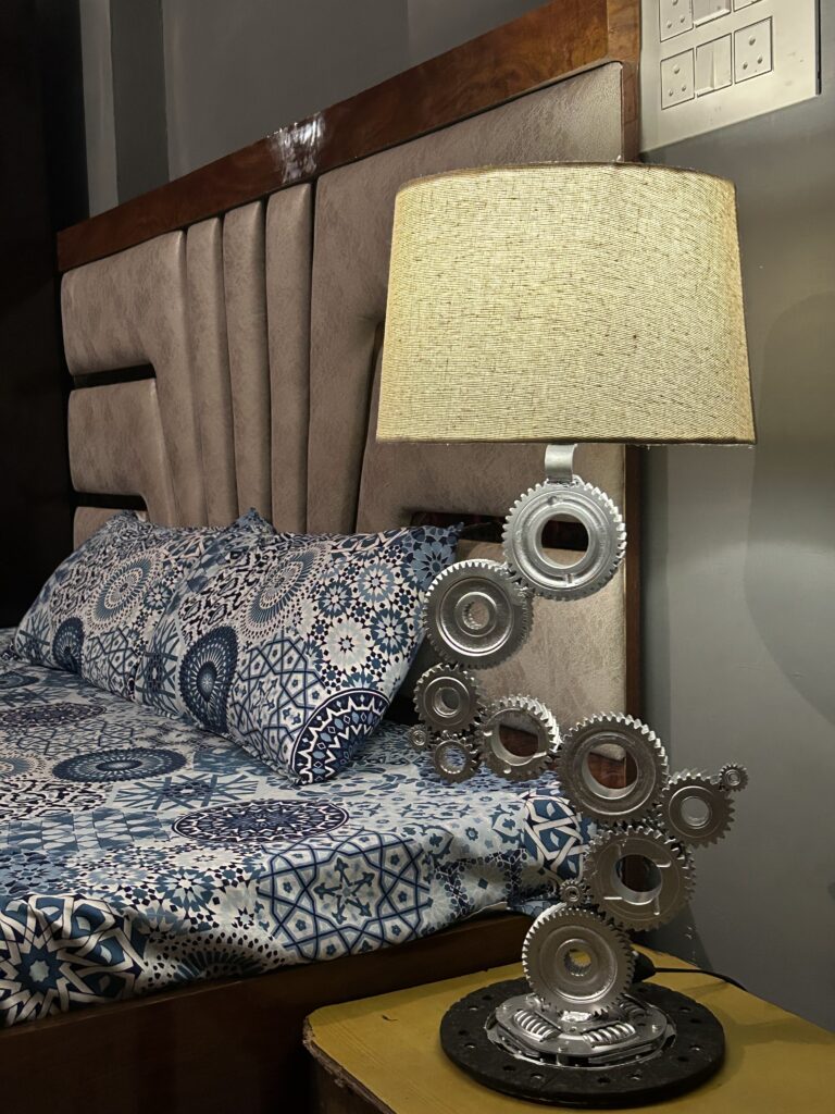 Handcrafted industrial automotive gear lamp sitting on a bedroom nightstand next to a bed. Unique upcycled car part gift for husband or boyfriend.